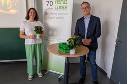 Zwei Personen stehen in einem Raum nebeneinander. Die eine Person hält einen kleinen Blumenstrauß in der Hand. Zwischen ihnen steht ein runder Tisch, auf dem ebenfalls eine Pflanze platziert ist. Im Hintergrund ist ein Roll-Up-Banner mit der Aufschrift „70 Jahre rehawiss – Der Blick zurück nach vorn“ zu sehen, darunter die Ergänzung „Fakultät Rehabilitationswissenschaften“.
