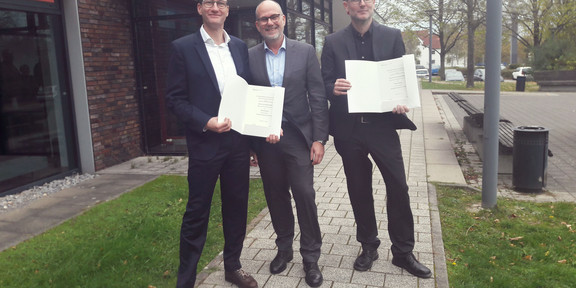 Drei Personen in schwarzen Anzügen (von links nach rechts: Vetr.-Prof. Dr. Bastian Pelka, Prof. Dr. Jan Kuhl und Dr. Christoph Kaletka) stehen auf dem gepflasterten Gehweg vor einem Gebäude. Die beiden äußeren Personen halten jeweils eine Urkunde in der Hand.