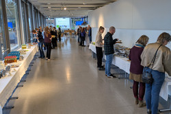 In einem hellen Flur mit großen Fenstern sind lange Buffet-Tische aufgebaut. Menschen stehen in kleinen Gruppen zusammen oder bedienen sich am Buffet. 