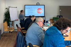 Das Bild zeigt einen modernen Schulungsraum, in dem eine Präsentation stattfindet. An einem langen Tisch sitzen mehrere Teilnehmer,*innen die auf den Bildschirm vor ihnen schauen. Der Bildschirm zeigt das Thema „Fragebogen 1.1“ und ein Stoppschild-Symbol, was möglicherweise auf eine wichtige Anweisung hinweist. Eine Person steht vorne im Raum und erläutert den Inhalt der Präsentation.