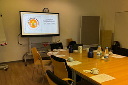 Das Bild zeigt einen Konferenzraum, der für eine Besprechung vorbereitet ist. An einem großen Tisch stehen Stühle, die mit Geschirr und Getränken gedeckt sind. Auf dem Tisch befinden sich Kaffeetassen, Gläser und Wasserflaschen sowie eine Kanne. An einer Wand hängt ein großer Bildschirm, auf dem das Thema „Umgang mit Schönheitsidealen auf Social Media“ angezeigt wird. 
