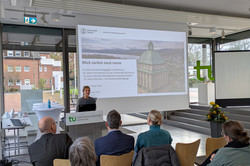 Ein Vortrag findet in einem hell beleuchteten Raum statt. Eine Person steht am Rednerpult. Mehrere Personen sitzen im Publikum und hören aufmerksam zu.