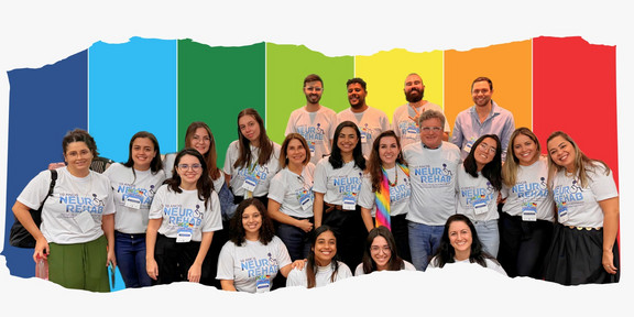 Zu sehen sind Studierende und Professor*innen des NeuroRehab (Forschungs- und Versorgungszentrum für neuropsychomotorische Rehabilitation) vor einem Regenbogenhintergrund, aufgenommen im Rahmen des internationalen Kongresses zum zehnjährigen Bestehen des Zentrums in Brasilien.
