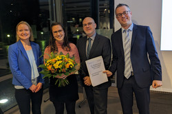 Von Links nach rechts stehen Prof. Dr. Janin Brandenburg, Lotte-Kaliski-Preisträgerin Marcia Klinger, Prof. Dr. Jan Kuhl und Prof. Dr. Jörg-Tobias Kuhn nebeneinander. Sie lächeln in die Kamera. Marcia Klinger hält einen Blumenstrauß in den Händen.