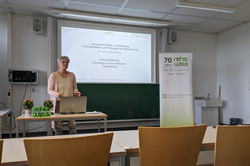 Eine Frau mit grauen, kurzen Haaren, Brille und heller Kleidung steht in einem Seminarraum hinter einem Tisch und spricht vor einer Projektionsleinwand. Auf der Leinwand ist der Titel des Vortrags zu lesen: „Fachgebiet Musik und Bewegung in Rehabilitation und Pädagogik bei Behinderung – Kulturelle Bildung: Unterwegs zu einer inklusiven Gesellschaft“. 