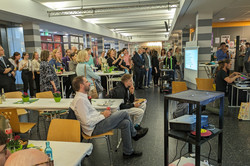 Eine größere Menschenmenge steht und sitzt im Foyer des EF50 und verfolgt eine musikalische Aufführung. Es gibt Stehtische mit grüner Deko und Getränken. Rechts sitzt eine Person an einem Mischpult. Im Hintergrund ist eine Projektion mit Liedtexten sichtbar.