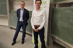 Zwei Personen stehen in einem Vorlesungssaal vor einer Tafel. Eine Person trägt einen dunklen Blazer mit Jeans, die andere ein weißes Hemd und dunkelblaue Hosen mit auffälligen roten Sneakern. Hinter ihnen ist ein Roll-Up-Banner mit der Aufschrift „70 Jahre rehawiss – Der Blick zurück nach vorn“ zu sehen, darunter die Ergänzung „Fakultät Rehabilitationswissenschaften“. Im Hintergrund ist eine Projektion an der Wand sichtbar, auf der „Rehabilitationssoziologie“ und „Bastian Pelka“ steht.