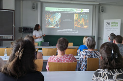 Eine Person steht vorne in einem Seminarraum und hält eine Präsentation. Auf der Leinwand ist eine Folie mit der Überschrift „Lösungsansätze DiFiB“ zu sehen, darunter der Text „durch eine gezielte universitäre Ausbildung zur digitalen Bildung“. Die Folie zeigt zwei Bilder: links eine Darstellung von Personen in einer Lichtszene und rechts ein Bild von Menschen in einem Klassenzimmer. 