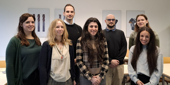 Eine Gruppe von sieben Personen steht in einem hellen Besprechungsraum. Sie posieren für ein Gruppenfoto vor einer weißen Wand.