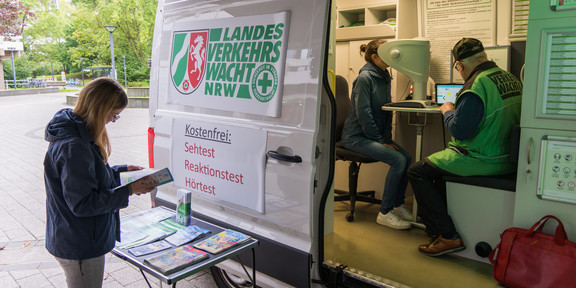 Eine Frau steht vor dem Sehtest-Mobil der Verkehrswacht NRW. Drinnen führt ein Mitarbeiter einen Sehtest mit anderen Frau durch.