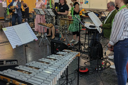 Die Musikgruppe der musikalischen Mittagspause spielt und singt im Foyer des EF50. Vorne im Bild steht ein großes Xylophon, daneben ein Notenständer mit Notenblättern. Die Musiker*innen stehen in einer Reihe, einige mit Mikrofonen, andere mit Percussion-Instrumenten. Ein Mann spielt Gitarre.