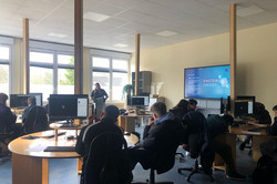 Ein Computerraum mit mehreren Schüler*innen, die an runden Tischen mit Monitoren sitzen. Eine Frau steht vorne neben einem großen Bildschirm, auf dem eine Präsentation mit dem Titel „Schutzschild Strategien“ läuft. 
