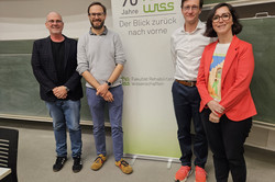 Vier Personen stehen nebeneinander in einem Seminarraum vor einer Tafel. Hinter ihnen ist ein Roll-Up-Banner mit der Aufschrift „70 Jahre rehawiss – Der Blick zurück nach vorn“ zu sehen, darunter die Ergänzung „Fakultät Rehabilitationswissenschaften“. Die Personen tragen unterschiedliche Kleidung, darunter Blazer, Pullover und farbige Sneaker.