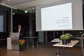 Prof. Dr. Manfred Bayer, Rektor der TU Dortmund, steht hinter einem Rednerpult. Hinter ihm ist eine Leinwand auf die das Wort "Grußwort" projiziert ist.