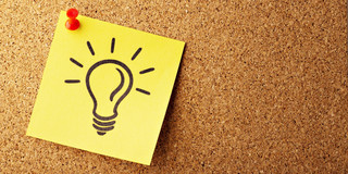 Auf einem quadratischen, gelben Post-it Zettel ist mit Edding eine Glühbirne gemalt. Der Post-It ist mit einer roten Stecknadel an einer Wand aus braunem Kork befestigt.