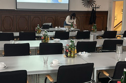 Das Bild zeigt einen Konferenzraum, der für eine Veranstaltung vorbereitet ist. In der Mitte des Raumes stehen mehrere lange Tische, die mit Getränken und Geschirr gedeckt sind. Auf den Tischen befinden sich Flaschen mit Wasser und Erfrischungsgetränken sowie Gläser und Tassen. An einer Wand hängt ein Projektorbildschirm, auf dem das Thema „Umgang mit sensiblen Inhalten auf Social Media“ angezeigt wird. Eine Person bereitet im Hintergrund den Raum vor.