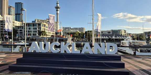 Ein großes Schild mit der Aufschrift „Auckland“, das sich im Hafen der Stadt befindet und hinter dem die Skyline zu erkennen ist.