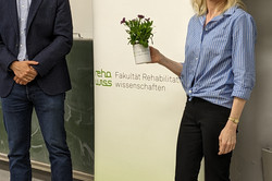 Ein Mann und eine Frau stehen in einem Vorlesungssaal neben einem Roll-up mit der Aufschrift „70 Jahre rehawiss – Der Blick zurück nach vorne – Fakultät Rehabilitationswissenschaften“. Der Mann links trägt eine Brille, ein dunkelblaues Sakko, ein hellblaues Hemd und Jeans. Die Frau rechts hat blondes, schulterlanges Haar und trägt eine blau-weiß gestreifte Bluse, schwarze Hose und braune Schuhe mit Absatz. Sie hält eine kleine Pflanze in einer weißen Tasse mit der Aufschrift der Veranstaltung. Beide lächeln in die Kamera.