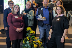 Eine Gruppe von Personen steht auf einer Treppe in einem Gebäude. Vor ihnen steht ein hoher schwarzer Blumenkübel mit gelben und roten Blumen. Im Hintergrund ist eine große, farbenfrohe Präsentationswand mit Texten und Illustrationen zu sehen.