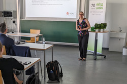 Eine Person steht in einem Seminarraum und hält eine Präsentation. Auf der Leinwand ist eine Folie mit dem Titel „Bildungsmonitoring“ zu sehen, die Informationen über Forschungsansätze und internationale Studien enthält, darunter die IEA (International Association for the Evaluation of Educational Achievement). Im Raum sitzen mehrere Personen an Tischen und folgen der Präsentation.