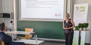 Eine Person steht in einem Seminarraum und hält eine Präsentation. Auf der Leinwand ist eine Folie mit dem Titel „Bildungsmonitoring“ zu sehen, die Informationen über Forschungsansätze und internationale Studien enthält, darunter die IEA (International Association for the Evaluation of Educational Achievement). Im Raum sitzen mehrere Personen an Tischen und folgen der Präsentation.