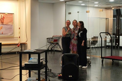 Auf dem Bild sind drei Frauen zu sehen, die Mikrofone in den Händen halten und Karaoke singen. Im Vordergrund ist ein kleiner Tisch mit einem Projektor und anderen Geräten zu sehen, während ein Lautsprecher in der Mitte des Raumes steht.