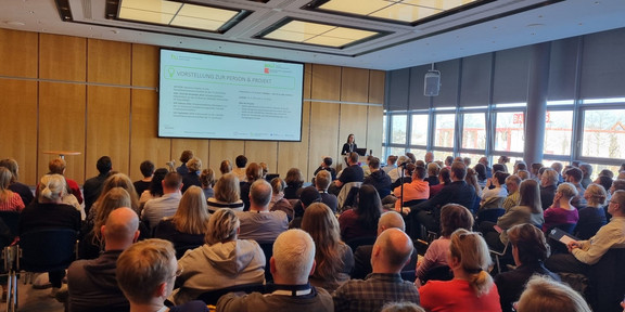 In einem Konferenzraum findet eine Präsentation statt. Im Vordergrund sieht man die Rückenansicht der Teilnehmer*innen, die in Reihen auf Stühlen sitzen und nach vorne blicken. Im Hintergrund steht eine Referentin an einem Pult neben einer großen Leinwand.