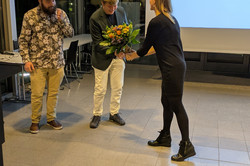 Eine Person in schwarzer Kleidung überreicht einem anderen Menschen einen Blumenstrauß. Neben ihnen steht eine weitere Person, die das Geschehen beobachtet.