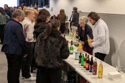 Ein heller Innenraum mit vielen Menschen, die sich an einem Getränkestand versammeln. Auf den Tischen stehen verschiedene Flaschen mit alkoholfreien und alkoholischen Getränken.