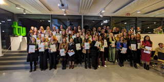 Gruppenfoto Absolvierendenfeier Alle anwesenden Absolventinnen und Absolventen stehen für ein Gruppenfoto bereit und lächeln in die Kamera. Sie halten einen weißen A4 Umschlag und eine Blume in den Händen.