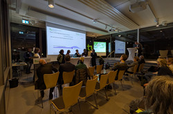 Im Veranstaltungsraum findet eine Podiumsdiskussion statt. Fünf Personen stehen oder sitzen an Stehtischen vor dem Publikum, während eine weitere Person am Rednerpult spricht. Auf der Leinwand ist eine Präsentationsfolie mit einer These zur Digitalisierung eingeblendet.