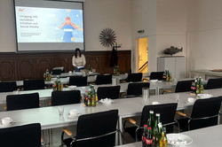 In dem Bild ist ein Konferenzraum zu sehen, der für eine Veranstaltung vorbereitet ist. In der Mitte des Raumes stehen mehrere lange Tische, die mit Getränken und Geschirr gedeckt sind. An einer Wand hängt ein Projektorbildschirm, auf dem eine Präsentation zum Thema "Umgang mit sensiblen Inhalten auf Social Media" angezeigt wird.
