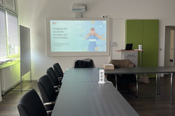 Das Bild zeigt einen modernen Schulungsraum, der für eine Präsentation vorbereitet ist. An einem langen Tisch stehen mehrere Stühle, die auf die Präsentation ausgerichtet sind. An einer Wand hängt ein Projektorbildschirm, auf dem das Thema „Umgang mit sensiblen Inhalten auf Social Media“ angezeigt wird.