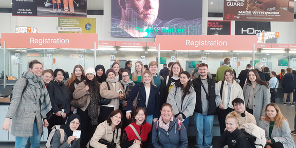 Gruppenfoto mit vielen Personen vor dem Eingang einer Messe. Im Hintergrund sind große Bildschirme mit Veranstaltungshinweisen zu sehen.
