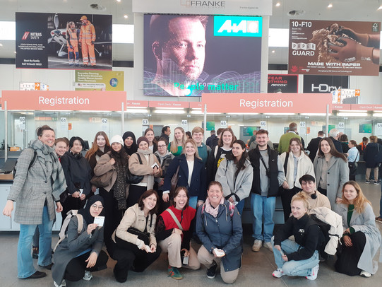 Gruppenfoto mit vielen Personen vor dem Eingang einer Messe. Im Hintergrund sind große Bildschirme mit Veranstaltungshinweisen zu sehen.