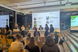 Auf dem Podium stehen fünf Diskutierende sowie zwei Moderierende an einem Rednerpult der TU Dortmund. Auf der Leinwand werden ihre Namen und Institutionen angezeigt.