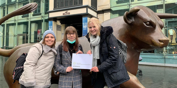 Studierende der Fakultät Rehabilitationswissenschaften stehen vor einer Statue in Birmingham und halten das Logo der Fakultät hoch