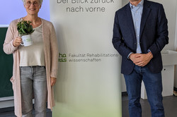 Eine Frau mit grauen, kurzen Haaren, Brille und heller Kleidung steht in einem Seminarraum hinter einem Tisch und spricht vor einer Projektionsleinwand. Auf der Leinwand ist der Titel des Vortrags zu lesen: „Fachgebiet Musik und Bewegung in Rehabilitation und Pädagogik bei Behinderung – Kulturelle Bildung: Unterwegs zu einer inklusiven Gesellschaft“.