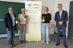 Vier Personen stehen nebeneinander vor einem Roll-up mit dem Schriftzug „70 Jahre rehawiss – Der Blick zurück nach vorne“. Zwei Männer und zwei Frauen. Alle halten weiße Becher mit kleinen Topfpflanzen. Von links nach rechts: Ein Mann mit dunkler Brille, Bart, blauem Sakko und Jeans. Eine Frau mit grauen, kurzen Haaren und Brille, heller Kleidung und grauen Schuhen. Eine blonde Frau mit Brille, schwarzem Oberteil und heller Jeans. Ein Mann mit Brille, dunkelblauem Sakko und Jeans, lächelt in die Kamera