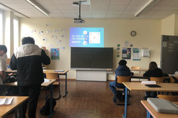 Ein Klassenzimmer mit mehreren Schüler*innen, die auf eine Präsentation schauen. Die Folie auf dem Beamer zeigt die Frage „Wie kann man sich vor Cybermobbing schützen?“. Einige Schüler*innen sitzen an Tischen, andere stehen. An der Wand hängen Sprechblasen mit positiven Botschaften und ein Plakat.
