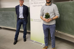 Zwei Personen stehen in einem Seminarraum vor einer Tafel. Eine Person trägt einen Pullover und hält eine kleine Pflanze in den Händen. Hinter ihnen ist ein Roll-Up-Banner mit der Aufschrift „70 Jahre rehawiss – Der Blick zurück nach vorn“ zu sehen, darunter die Ergänzung „Fakultät Rehabilitationswissenschaften“.
