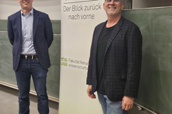 Zwei Personen stehen in einem Vorlesungssaal vor einer Tafel. Eine trägt einen dunklen Blazer mit Jeans, die andere ein kariertes Sakko mit schwarzem Shirt und Jeans. Hinter ihnen ist ein Roll-Up-Banner mit der Aufschrift „70 Jahre rehawiss – Der Blick zurück nach vorn“ zu sehen, darunter die Ergänzung „Fakultät Rehabilitationswissenschaften“. Im Hintergrund ist ein Teil einer Präsentation an der Wand erkennbar. 