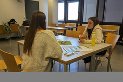 Zwei Personen führen ein Gespräch an einem kleinen Tisch in einem hellen Seminarraum.