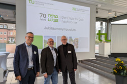 Drei Personen stehen nebeneinander vor einer großen Projektionsfläche mit dem Schriftzug „TU Dortmund – Fakultät Rehabilitationswissenschaften; Der Blick zurück nach vorne; Jubiläumssymposium“.