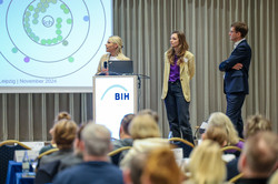 Ein blonde Frau steht auf einer Bühne während sie den Fachvortrag auf dem bundesweiten Netzwerktreffen der EAA hält. Im Hintergrund ist ein Ausschnitt der Präsentation zu sehen. Eine Frau mit langen braunen Haaren und ein Mann mit kurzen braunen Haaren stehen ebenfalls auf der Bühne und hören zu.