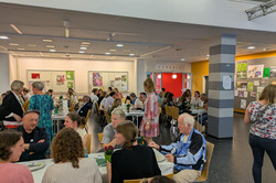 Das Bild zeigt das Foyer des EF50, in dem mehrere Personen an Tischen sitzen und sich unterhalten. Die Tische sind mit weißen Tischdecken und teilweise mit Dekoration wie Blumen und Getränken versehen. Einige Personen stehen im Hintergrund am Buffet, während die meisten sitzen, essen und sich unterhalten.