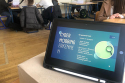 Das Bild zeigt einen Unterrichtsraum, in dem Schüler*innen an Tischen sitzen und an einer Gruppenarbeit teilnehmen. Im Vordergrund ist ein Tablet zu sehen, das die Anweisungen für die Gruppenarbeit zum Thema „Cybermobbing erkennen“ anzeigt. Die Aufgaben beinhalten die Bildung von vier Gruppen, wobei jede Gruppe ein Fallbeispiel erhält, das sie diskutieren soll.