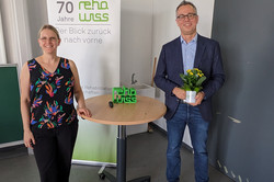 Ein Mann und eine Frau stehen nebeneinander an einem Tisch und schauen frontal in die Kamera. Der Mann hält eine Blume in der Hand. Beide lächeln freundlich.