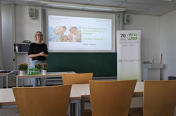 Eine blonde Frau mit Brille, schwarzem Oberteil und heller Jeans steht im Seminarraum vor einer Präsentationsleinwand. Auf der Leinwand ist der Vortragstitel zu sehen: „Das Fachgebiet Sehen, Sehbeeinträchtigung & Blindheit: Aktuelle Forschung“ von Sarah Weigelt. Der Vortrag ist Teil der Jubiläums-Ringvorlesung der Fakultät Rehabilitationswissenschaften an der TU Dortmund. Auf dem Tisch vor der Rednerin stehen zwei Blumentöpfe, ein Laptop und das grüne Logo „reha WISS“. 