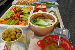 Auf dem Bild ist ein Buffet mit verschiedenen Speisen zu sehen, das auf einem Tisch angerichtet ist. Es gibt eine Vielzahl von Gemüse wie Möhren, Gurken und Tomaten sowie verschiedene Dips und Salate. 